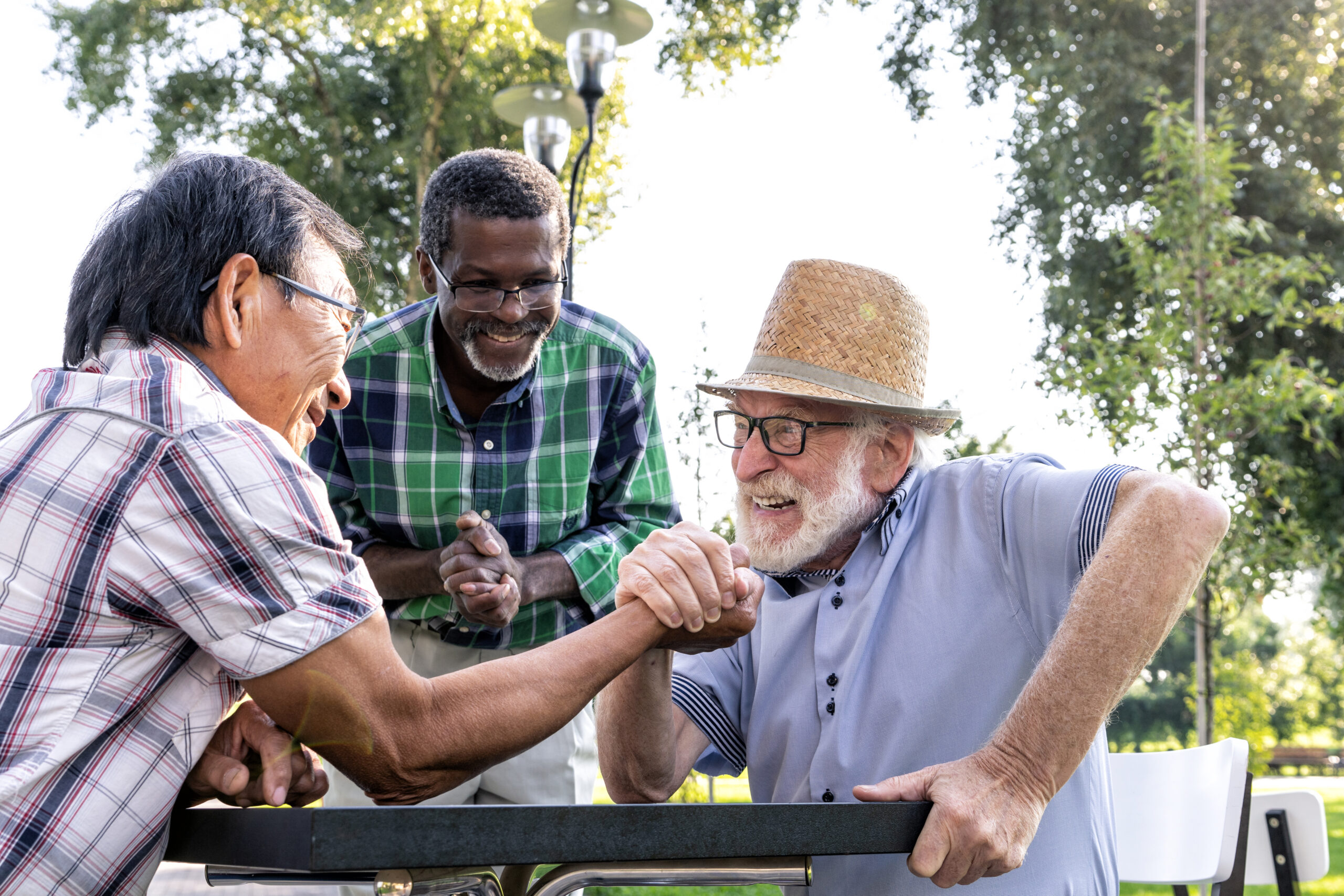 "Two elderly men arm wrestling playfully outdoors, supported by a smiling friend"