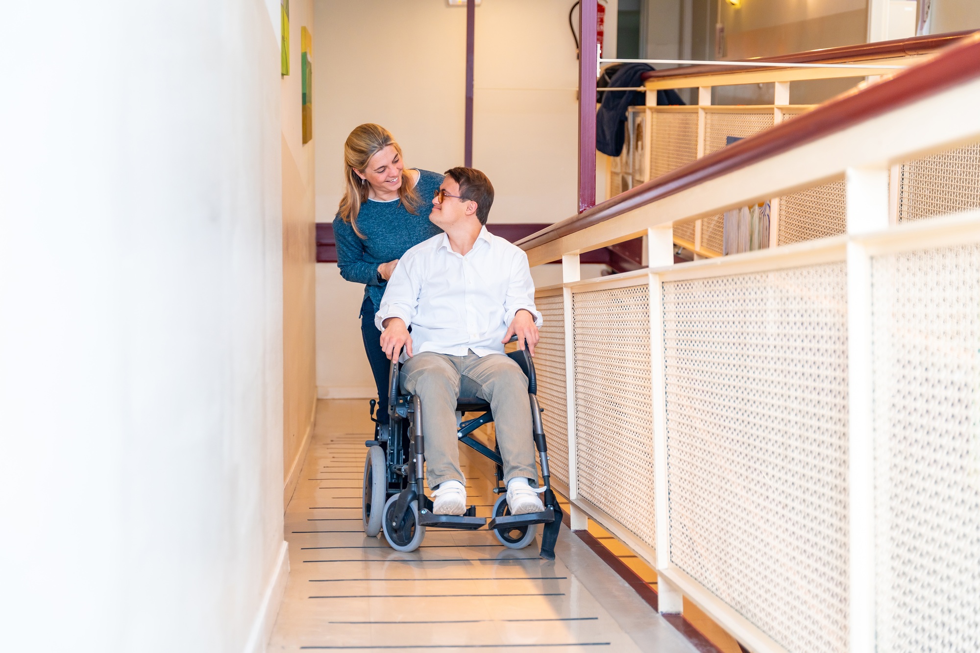 Smiling female nurse assisting a young man in a wheelchair along a hallway, showcasing community nursing support.